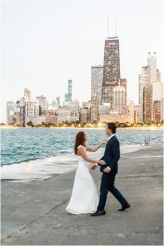 a bride and groom walking by the water in front of a city skyline at sunset