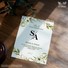 the wedding card is on top of a wooden table next to a candle and some greenery