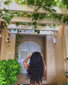 a woman with long hair standing in front of a doorway surrounded by plants and greenery