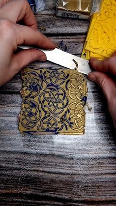 two hands holding scissors over a piece of gold and blue paper on top of a wooden table