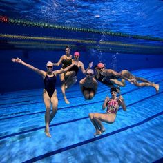 six people are swimming in an indoor pool and posing for the camera with their hands up