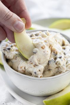 a hand dipping an apple into a bowl filled with cream cheese and chocolate chip cookies