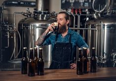 a man standing in front of a table filled with beer bottles drinking from a mug