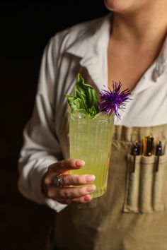 a woman in an apron holding a green drink with purple flowers on the top and leaves sticking out of it