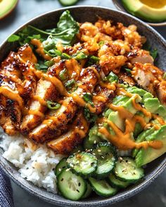 a bowl filled with rice, cucumber and chicken covered in sauce next to sliced avocado