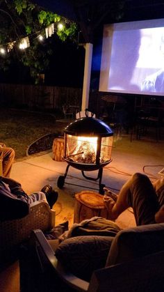 two people sitting in chairs watching a movie on the screen outside at night with an open fire pit