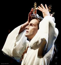 a man with long hair wearing a crown and holding his hands up in the air