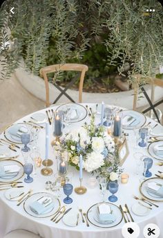 a white table topped with lots of blue and gold place settings next to tall plants