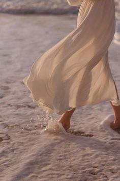 a woman in a white dress walking on the beach with her feet in the sand