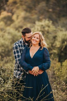 a man and woman standing in tall grass with their arms around each other, smiling at the camera