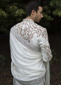 a man wearing a white suit with gold embroidery on the chest and back, standing in front of trees