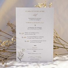 a white and gold wedding card on top of snow covered ground with dried flowers in the background