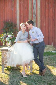 a man and woman sitting on a bench in front of a red barn with flowers