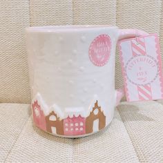 a white coffee mug sitting on top of a couch next to a pink and white tag