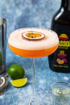 an orange cocktail in a coupe glass with limes and a bottle next to it