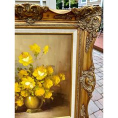 a painting with yellow flowers in a gold vase on a brick sidewalk next to a building