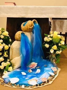 a teddy bear sitting on top of a table next to some flowers and shells in front of it