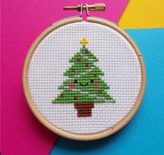 a cross stitch christmas tree on a white hoop hanging from a pink and blue background