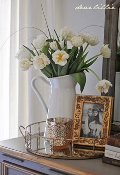 a white pitcher with flowers in it sitting on top of a table next to a mirror