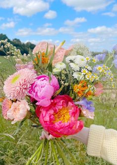 a person holding a bouquet of flowers in their hand on the grass with blue sky and clouds behind them