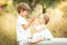 two young children playing with each other on a bench in the grass and trees behind them