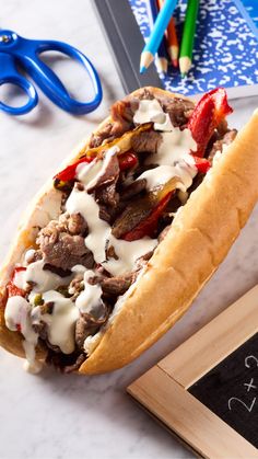 a sandwich with meat, cheese and peppers sitting on a table next to some pencils