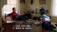 two women sitting on a bed in a room with posters above them and the words i like this song it makes me gloom