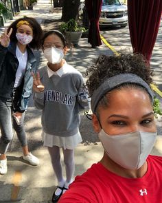 three girls wearing face masks and standing on the sidewalk with one pointing at the camera