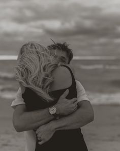 a man and woman hug on the beach while looking at the ocean in black and white