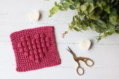 a crocheted dishcloth next to some scissors and flowers on a white table