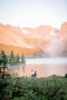 two people are sitting on the shore of a lake with mountains in the background and mist rising from the water