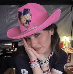 a woman wearing a pink cowboy hat and bracelets sitting at a table with her hand on her face