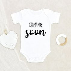 a white baby bodysuit with the words coming soon on it next to a heart