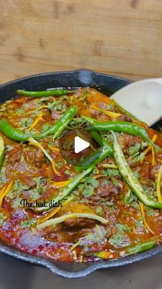 a skillet filled with meat and vegetables on top of a wooden table next to a spoon