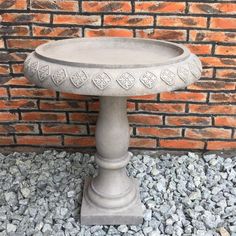 a cement bird bath sitting on gravel next to a brick wall and stone flooring