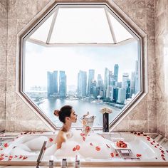 a woman sitting in a bathtub looking out the window at a cityscape