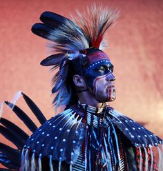 a native american man wearing an elaborate headdress and feathers on his face, standing in front of a red wall