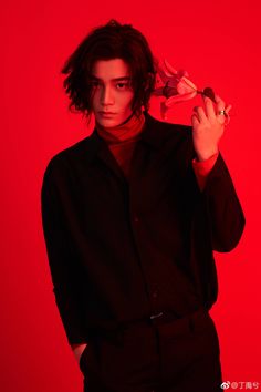 a young man in a black shirt and red background holding a flower up to his ear