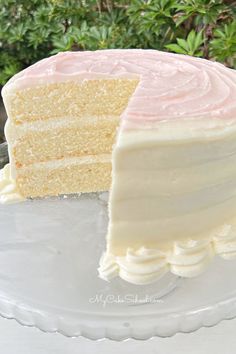 a white cake with pink frosting is on a glass platter next to some bushes