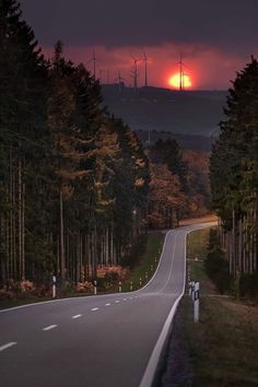 the sun is setting on an empty road in the woods with wind mills behind it