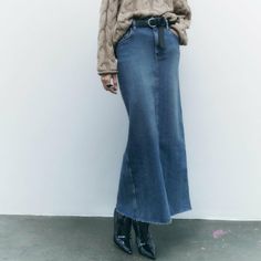 a woman standing in front of a white wall wearing a long denim skirt and sweater