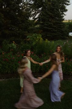 three women in long dresses holding hands and walking through the grass with flowers behind them