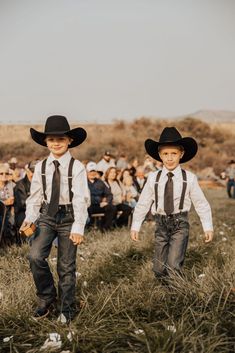 two young boys wearing cowboy hats and ties walk through the grass in front of an audience