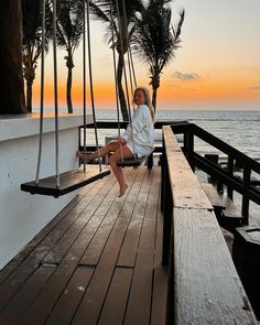 a woman sitting on a swing next to the ocean at sunset with palm trees in the background