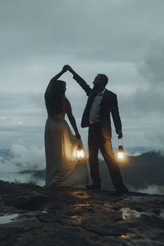 a man and woman standing on top of a mountain holding lanterns in their hands with clouds behind them