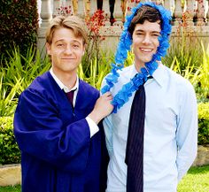 two men standing next to each other in front of a house wearing graduation gowns