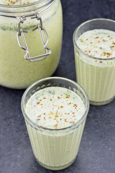 three glasses filled with green liquid sitting on top of a blue counter next to a jar