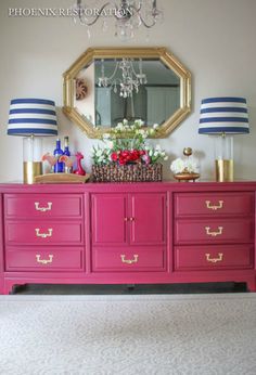 a pink dresser with blue and white striped lamps on top, and a gold framed mirror above it