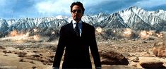 a man wearing a suit and tie standing in the desert with mountains in the background