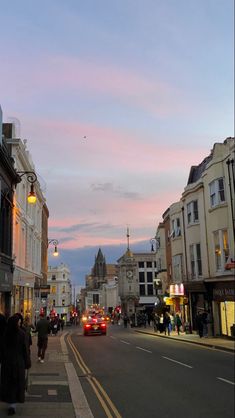 people are walking down the street in front of some buildings and cars at sunset or dawn